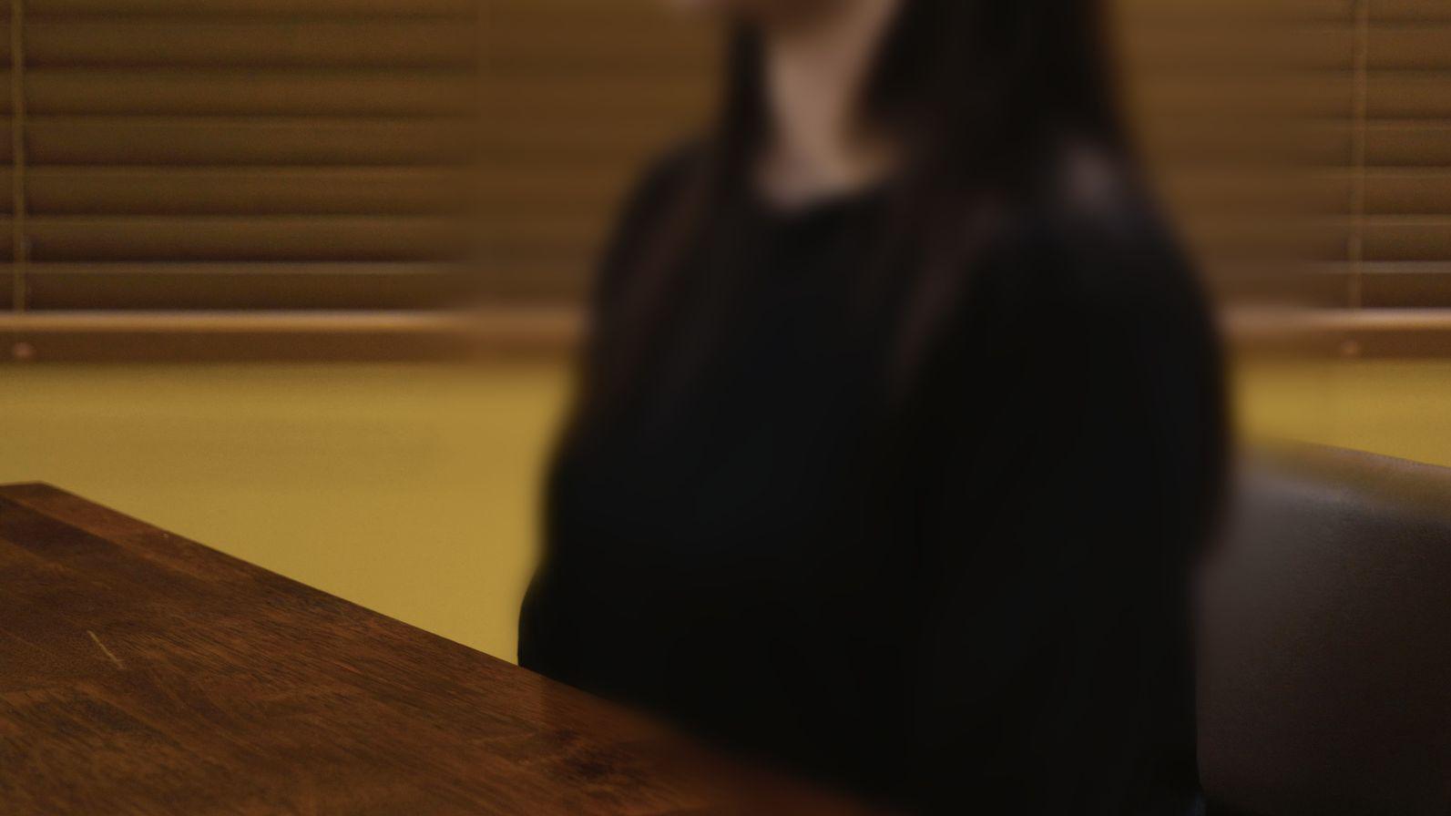 Una imagen borrosa de una mujer con una blusa negra, sentada detr&aacute;s de un escritorio.