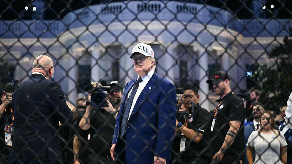 El presidente de los Estados Unidos, Donald Trump, visto a trav&eacute;s de una cerca de alambre met&aacute;lico. Lleva un traje azul y una gorra de b&eacute;isbol blanca que dice "EE. UU.". Al fondo hay una multitud de personas y la Casa Blanca