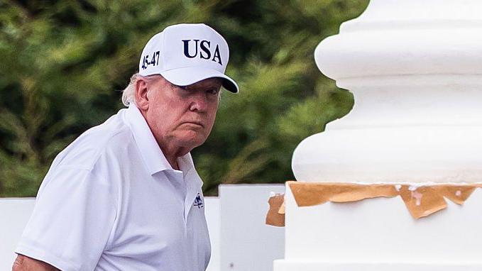 Donald Trump, vestido con polo blanco, pantal&oacute;n negro y una gorra blanca con el lema "USA", junto a una gran columna blanca. 