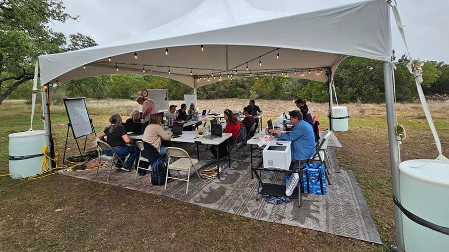 Un grupo de aproximadamente 12 personas se sienta en mesas ligeras trabajando con sus computadoras port&aacute;tiles en un campo, resguardadas bajo una gran tienda blanca con luces.