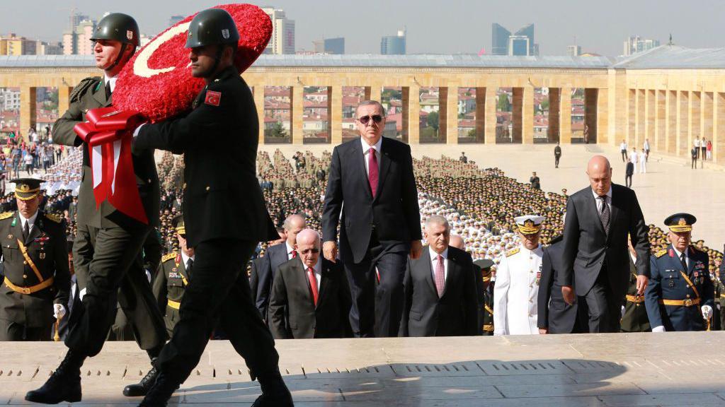 El presidente Recep Tayyip Erdogan sube los escalones del Mausoleo de Atat&uuml;rk en Ankara durante una ceremonia del D&iacute;a de la Victoria el 30 de agosto de 2016, flanqueado por funcionarios del gobierno mientras soldados portan una corona de flores con la forma de la bandera turca.