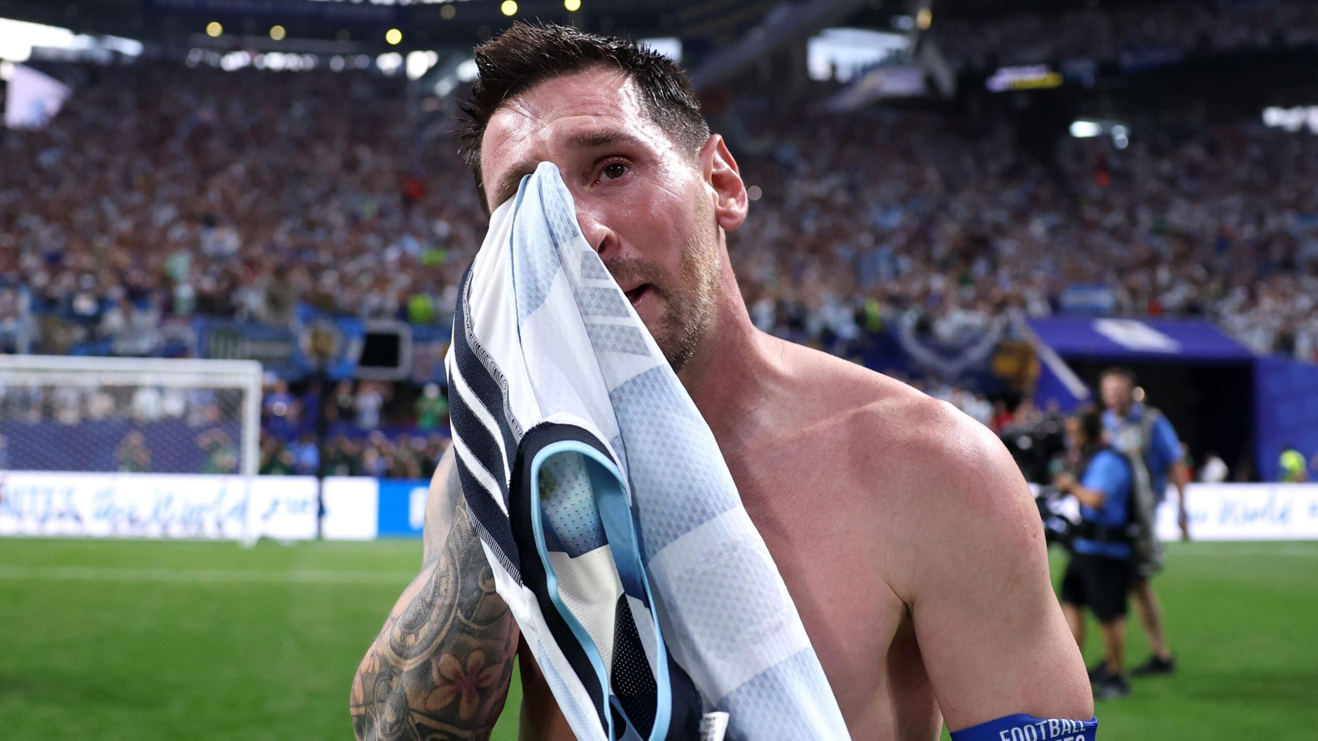 Lionel Messi en un estadio de f&uacute;tbol, junto al campo, sec&aacute;ndose las l&aacute;grimas del rostro con una camiseta mientras lleva un brazalete azul de la FIFA.