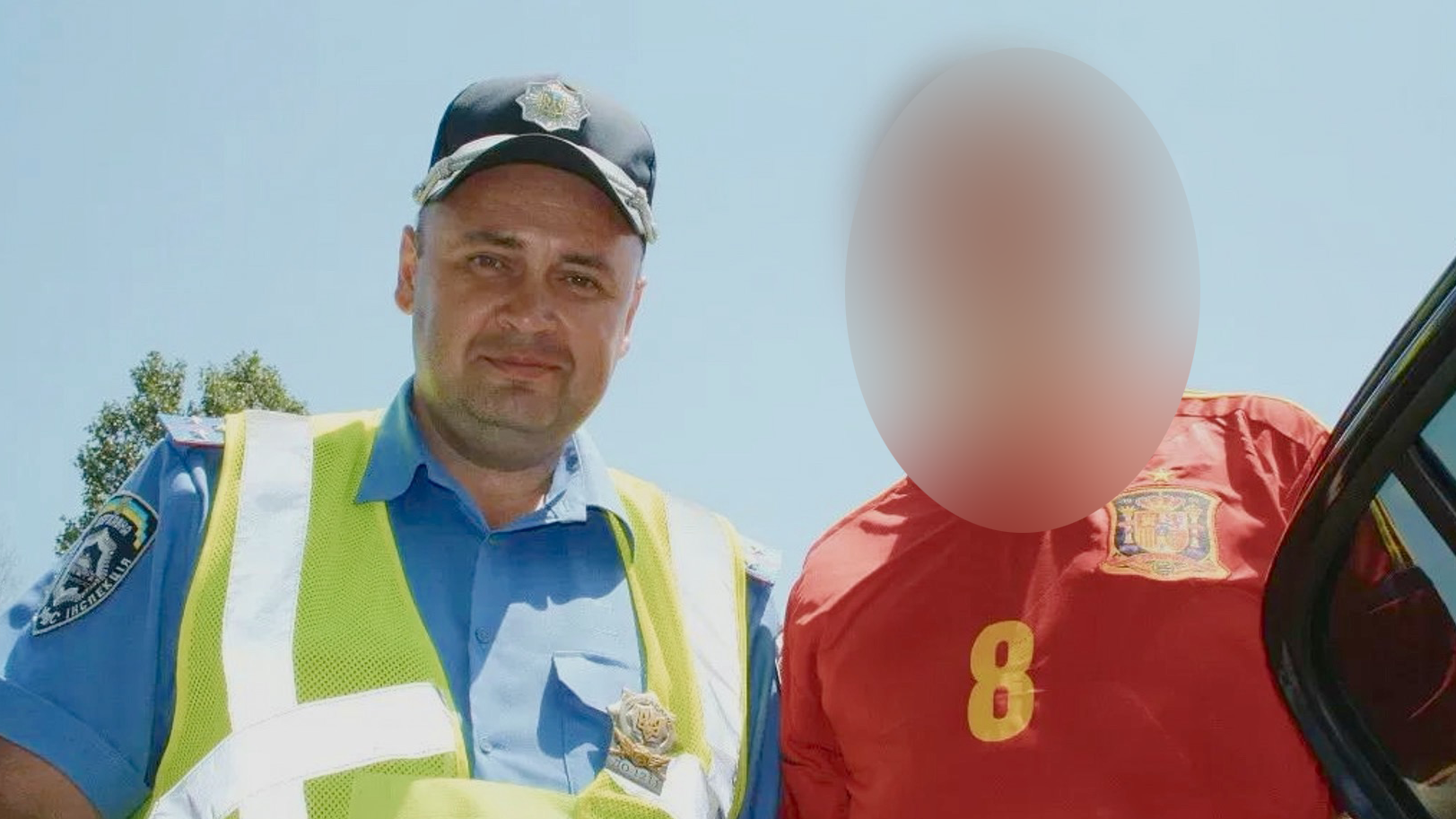 Temerbek aparece en la imagen con una gorra policial negra, una camisa de polic&iacute;a azul y un chaleco amarillo de alta visibilidad, mirando a la c&aacute;mara con una leve sonrisa. A su lado se encuentra una persona con una camiseta de f&uacute;tbol roja, cuyo rostro ha sido difuminado.