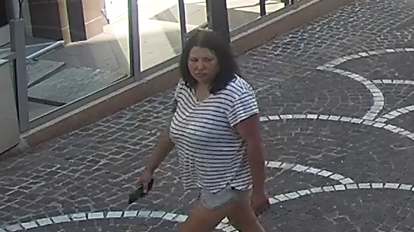 Una mujer de cabello oscuro y expresi&oacute;n seria mira hacia el otro lado de la calle por la que camina. Lleva una camisa a rayas blancas y negras. En una mano sostiene lo que parece ser un tel&eacute;fono m&oacute;vil.
