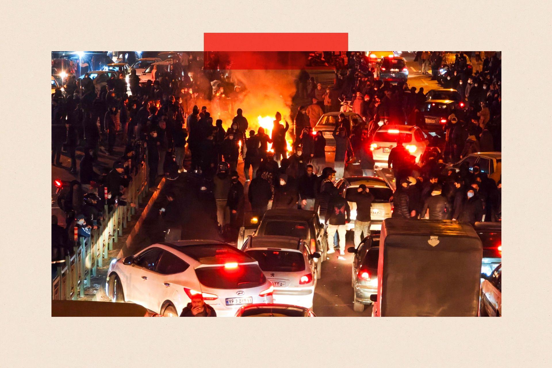 Cientos de personas protestando en las calles, con una hoguera a lo lejos y coches en primer plano.