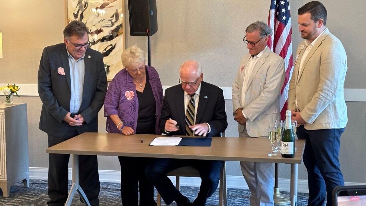 El gobernador de Minnesota, Tim Walz, con traje oscuro y corbata a rayas, ntras está sentado a una mesa con una carpeta abierta frente a él y sostiene un bolígrafo, preparándose para firmar una ley. Anita LeBrun, vestida con chaqueta morada, camisa negra y pantalones negros, está de pie junto a él. 