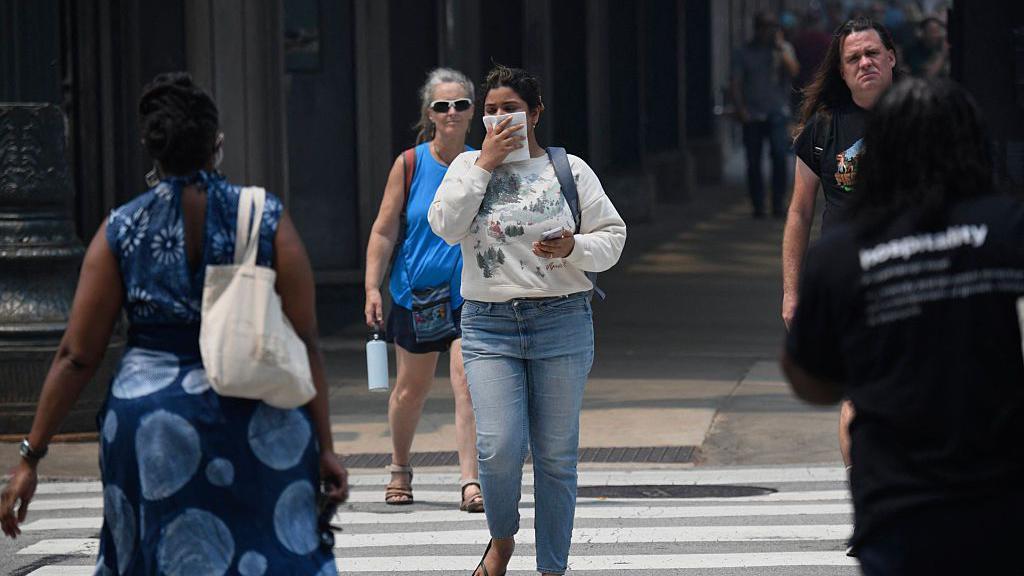 Una mujer vestida con una sudadera blanca y jeans se cubre el rostro con un pa&ntilde;uelo mientras cruza una calle de la ciudad, junto con otros peatones. 