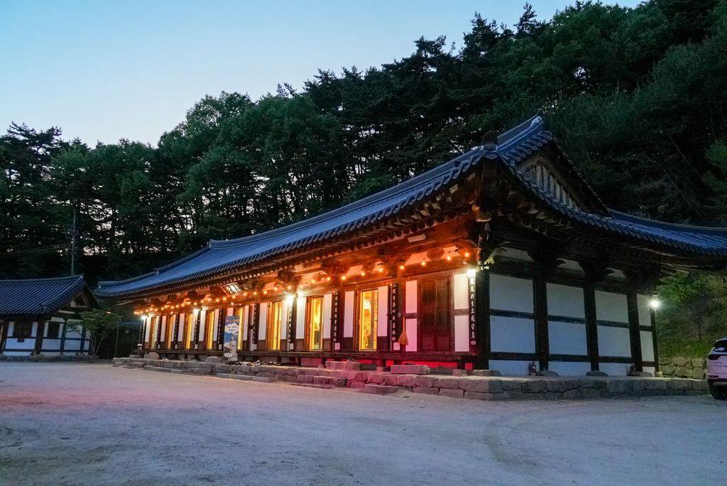 Templo tradicional coreano iluminado con luces c&aacute;lidas al atardecer, situado en un tranquilo patio al borde de una ladera boscosa.