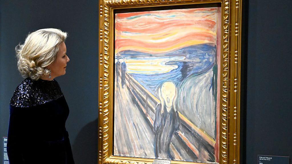 Un cuadro enmarcado que muestra a una persona gritando contra un cielo rojo brillante se exhibe en una galer&iacute;a, con un visitante de pie junto a &eacute;l contemplando la obra de arte.