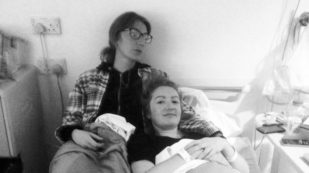 Fotograf&iacute;a en blanco y negro de Lowri en el hospital en septiembre de 2015, esperando unas pruebas junto a su hermana. Lowri est&aacute; acostada en la cama y su hermana la abraza. Ambas llevan el pelo recogido. La hermana de Lowri lleva una sudadera con capucha.
