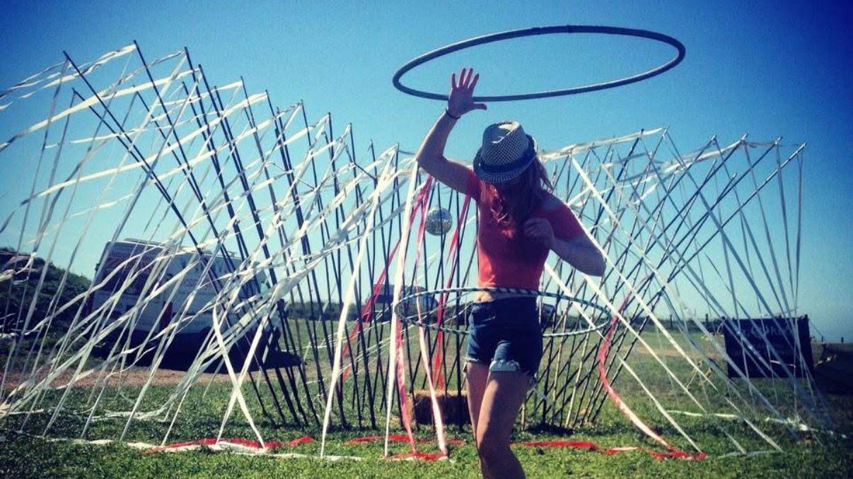 Lowri baila con un aro de hula-hula alrededor de la cintura. Lleva una blusa roja de manga corta y pantalones cortos vaqueros, y se encuentra en un campo con una instalaci&oacute;n art&iacute;stica cubierta de serpentinas detr&aacute;s de ella.