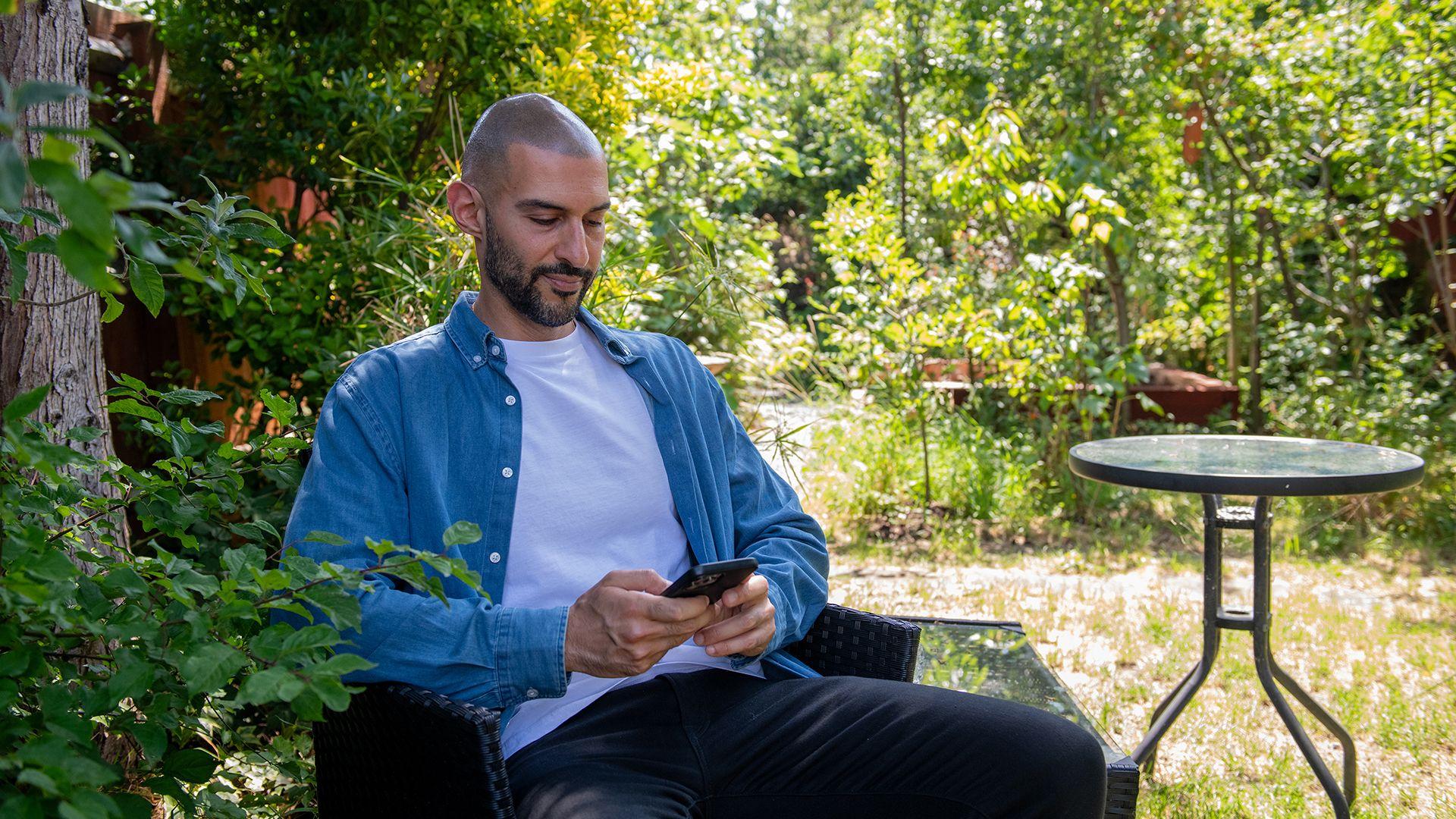 Un hombre con bigote y barba oscuros con camisa azul, camiseta blanca y pantalones negros, sentado en un jard&iacute;n, mirando su tel&eacute;fono m&oacute;vil. 