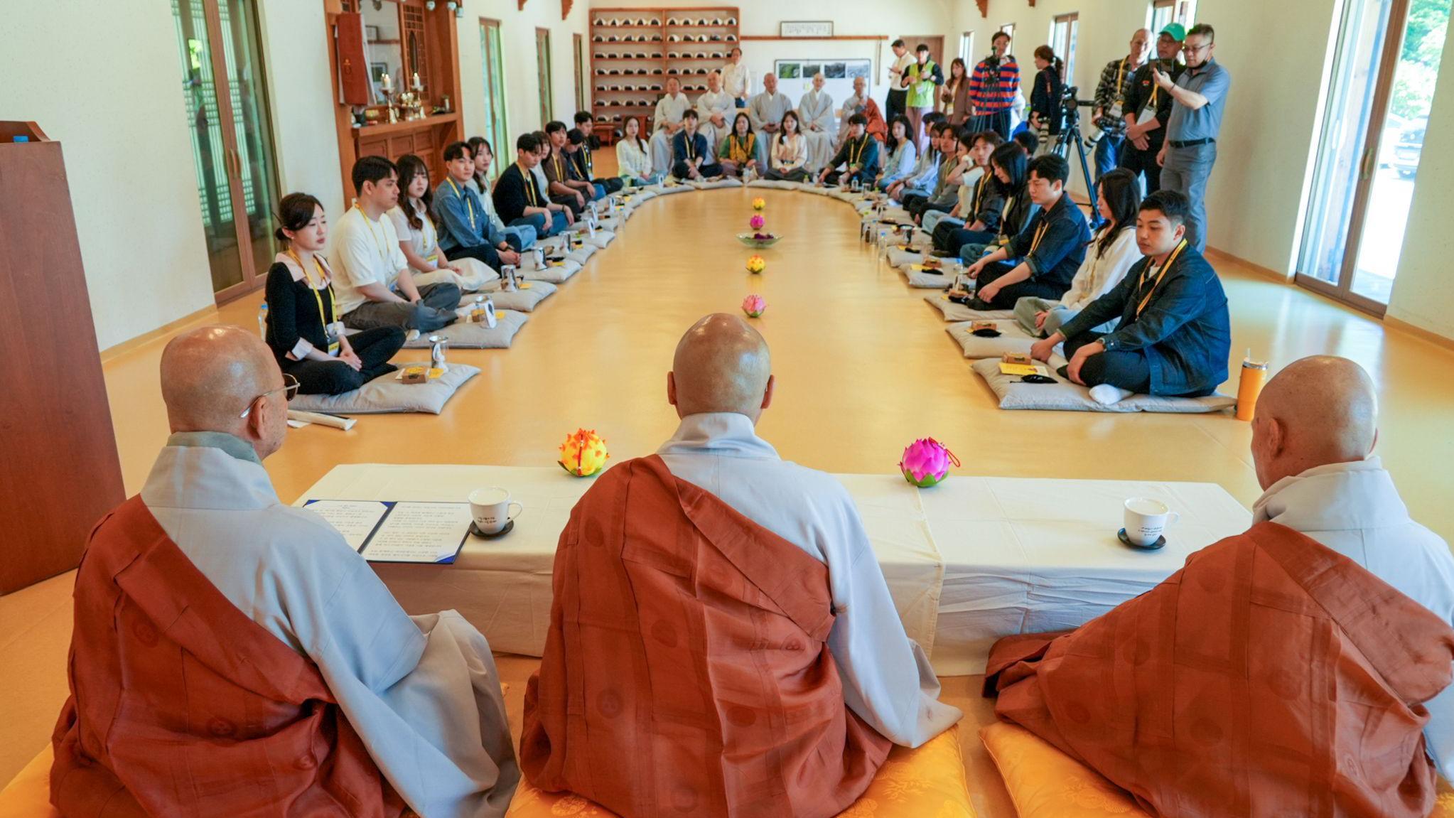 Un numeroso grupo de participantes est&aacute; sentado sobre cojines colocados en un amplio semic&iacute;rculo en el interior de la sala de un templo, con tres monjes al frente.