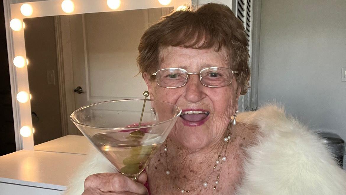 Lillian Droniak, de 96 años, sostiene una copa de martini con aceitunas. Sonríe mientras lleva puestas unas gafas, una estola de piel y un collar. Detrás de ella se ve un espejo enmarcado con luces.
