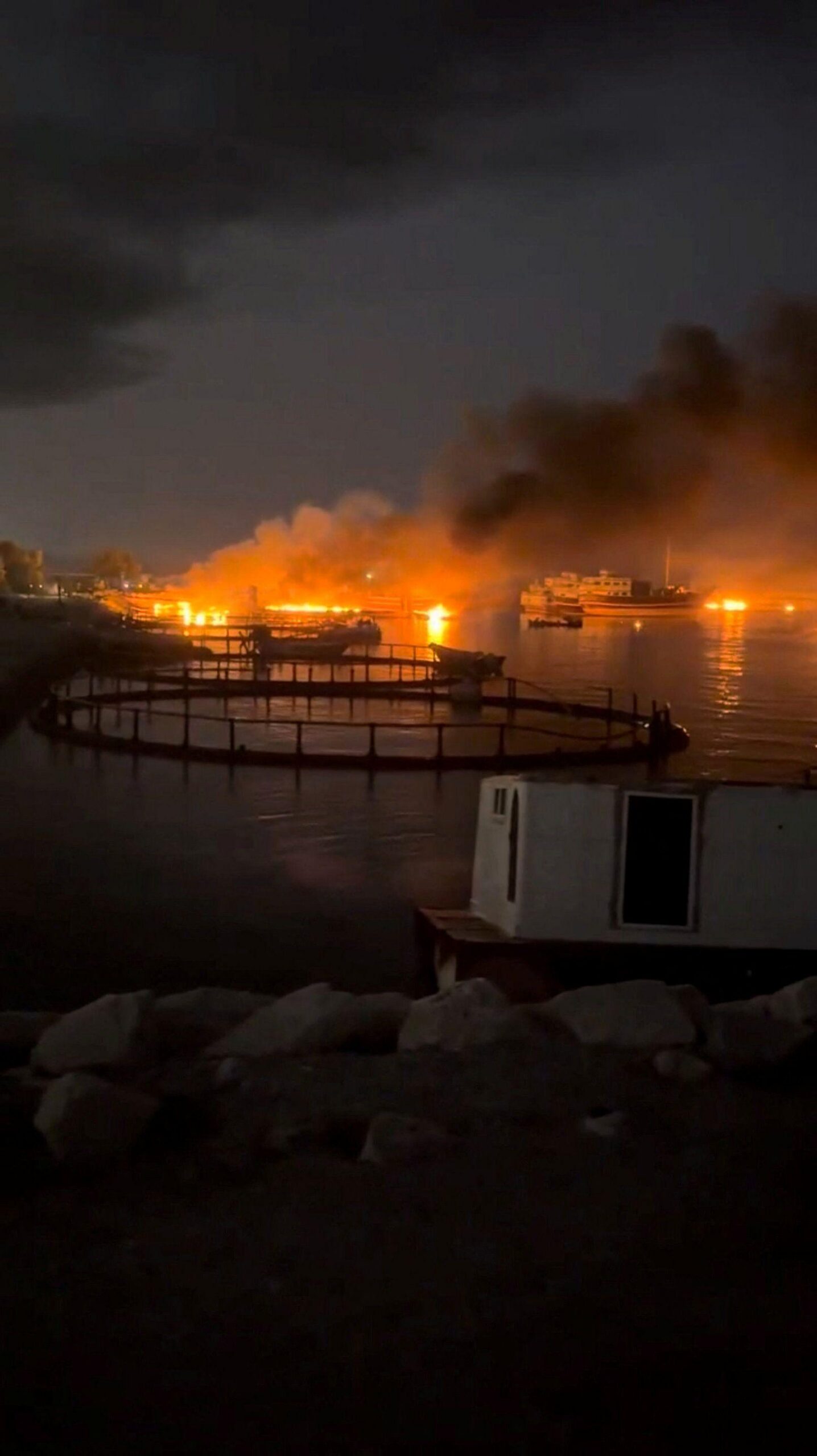 Vista nocturna de un incendio en el puerto. Llamas de color naranja brillante y una densa columna de humo negro se elevan desde varios puntos de la costa y se reflejan en la superficie del agua. Se aprecian varias embarcaciones cerca del fuego, mientras que en primer plano se distinguen estructuras marinas circulares y rocas.