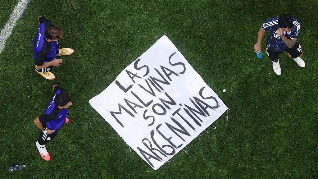 Una pancarta que dice "Las Malvinas Son Argentinas" est&aacute; en el suelo de un campo de juego con tres jugadores de pie a su alrededor.
