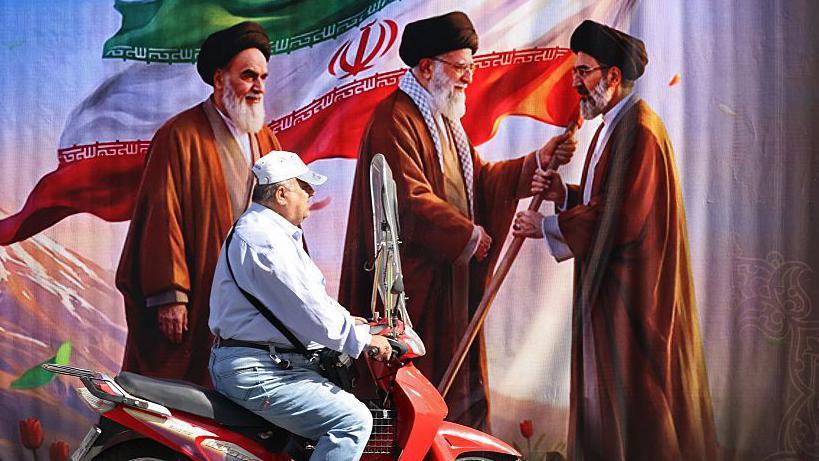 Un hombre circula en motocicleta frente a una valla publicitaria que muestra las im&aacute;genes del difunto fundador de la Revoluci&oacute;n Isl&aacute;mica y L&iacute;der Supremo, el ayatol&aacute; Ruhollah Jomeini (izquierda); del fallecido L&iacute;der Supremo de Ir&aacute;n, Ali Jamenei (centro); y de su hijo, el actual L&iacute;der Supremo, Mojtaba Jamenei, en una calle de Teher&aacute;n el 3 de julio de 2026