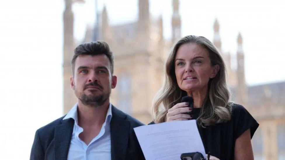 Con el parlamento de fondo, un hombre joven con barba y una mujer rubia sosteniendo un micr&oacute;fono y una hoja de papel.