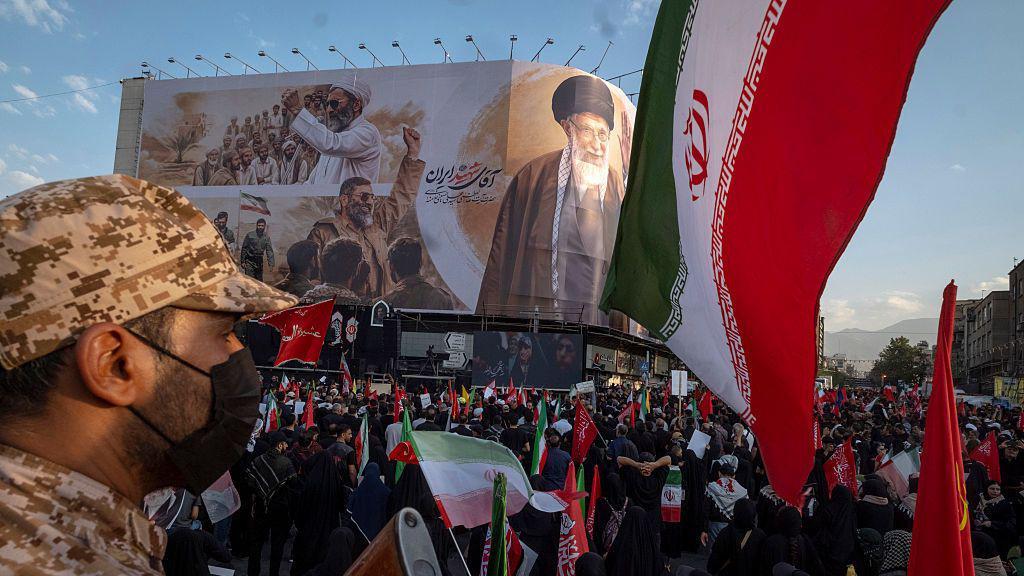 Un agente de seguridad iran&iacute; entre la multitud frente a una valla publicitaria que muestra al difunto l&iacute;der supremo en su funeral.