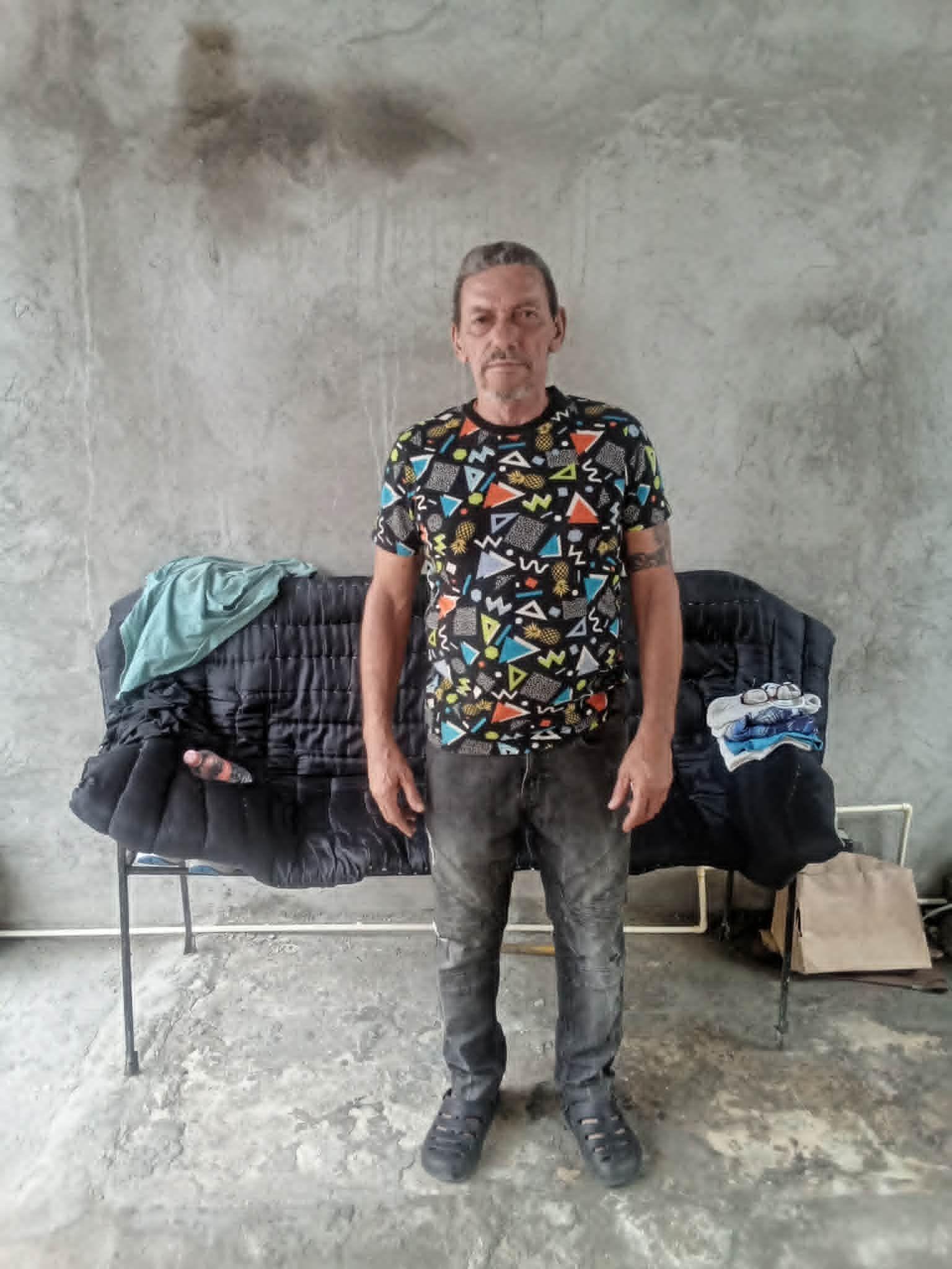 L&aacute;zaro Romero Le&oacute;n mira a c&aacute;mara de pie frente a una silla alargada, con jeans gris oscuros y una camiseta estampada, en un patio de cemento en Tapachula, Chiapas, M&eacute;xico.