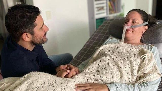 Paola Rold&aacute;n, incorporada en una cama, con un respirador en la nariz, tapada con una manta y agarrada de la mano de su esposo, a la izquierda y de perfil. 