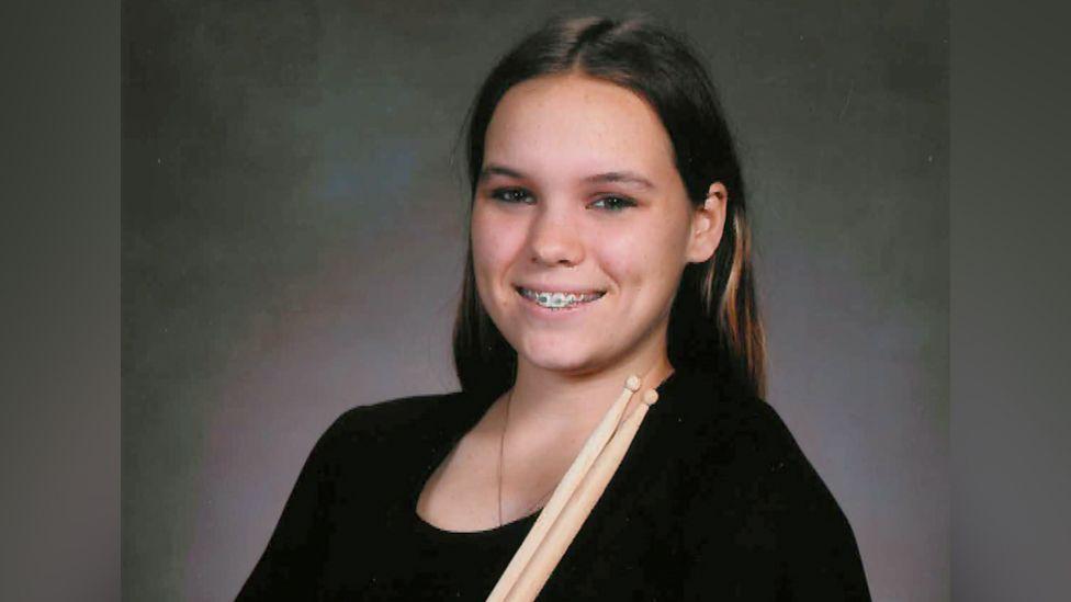 Taylor, a los 14 años, posa para un retrato. Viste una sencilla blusa negra de manga corta y sostiene dos baquetas de madera cruzadas sobre el pecho, mientras sorníe y en sus dientes se ven los aparatos que usaba en sus dientes.