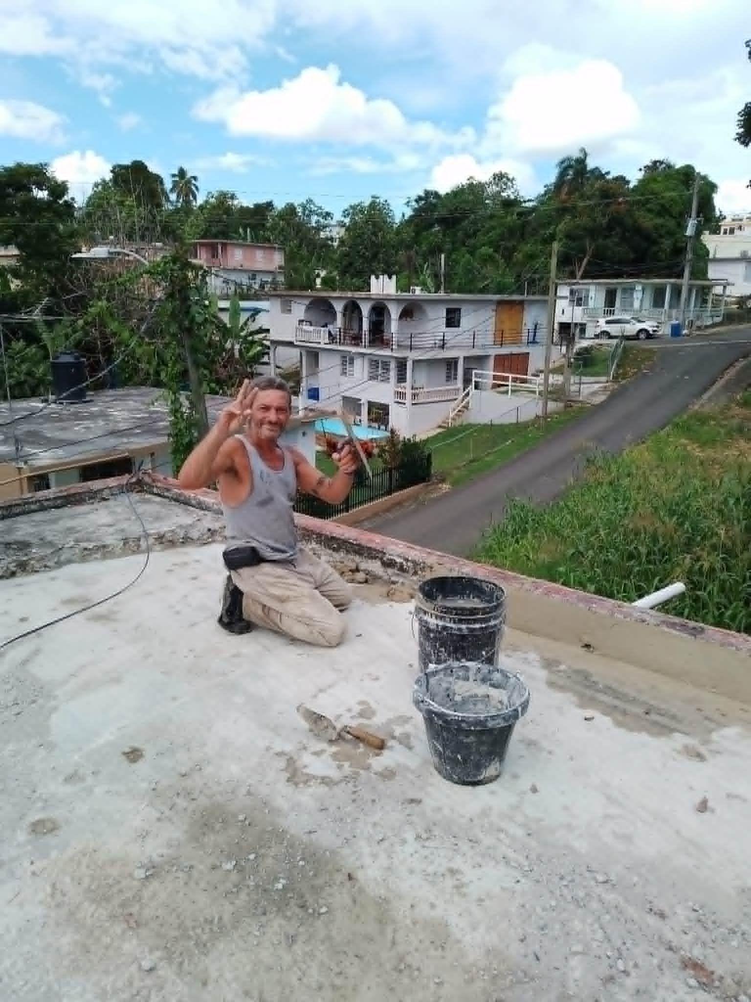 L&aacute;zaro Romero Le&oacute;n saluda a c&aacute;mara haciendo el gesto de la victoria con la mano derecha y una paleta en la izquierda, sobre el techo de una vivienda, junto a unos cubos con cemento, en Puerto Rico.