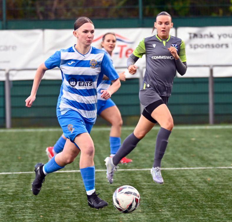 Tres mujeres jugando al f&uacute;tbol. Una mujer con una camiseta de rayas azules est&aacute; driblando el bal&oacute;n con una jugadora con una equipaci&oacute;n diferente cerca. 