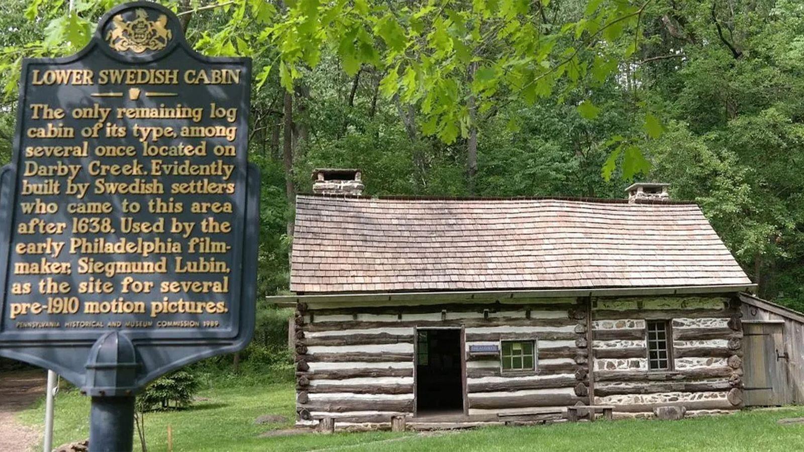 Caba&ntilde;a de troncos de origen sueco, conocida como Lower Swedish Cabin, una de las construcciones m&aacute;s antiguas de su tipo en Estados Unidos, junto a un cartel hist&oacute;rico en un entorno boscoso.
