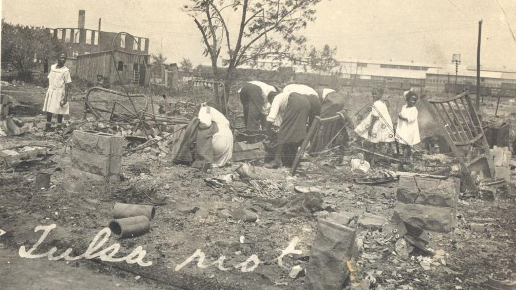 Una vieja fotograf&iacute;a en blanco y negro muestra a cinco adultos agachados, buscando entre los restos carbonizados de un edificio completamente destruido. Solo quedan algunos ladrillos y trozos de metal retorcido. A la derecha, dos ni&ntilde;as peque&ntilde;as con vestidos blancos observan la escena. A la izquierda, una mujer tambi&eacute;n observa. Al fondo se ve la estructura destruida de un edificio y, en la parte inferior de la foto, se lee "Disturbios de Tulsa" escrito con tinta blanca en una tipograf&iacute;a antigua.