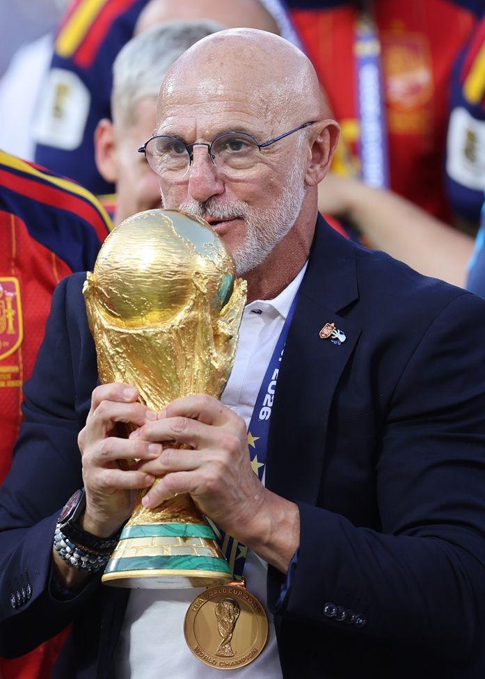 Luis de la Fuente, con lentes, camisa blanca y chaqueta azul oscura, carga el trofeo del Mundial con las dos manos. Est&aacute; rodeado de futbolistas espa&ntilde;oles. En su cuello, cuelga la medalla de oro