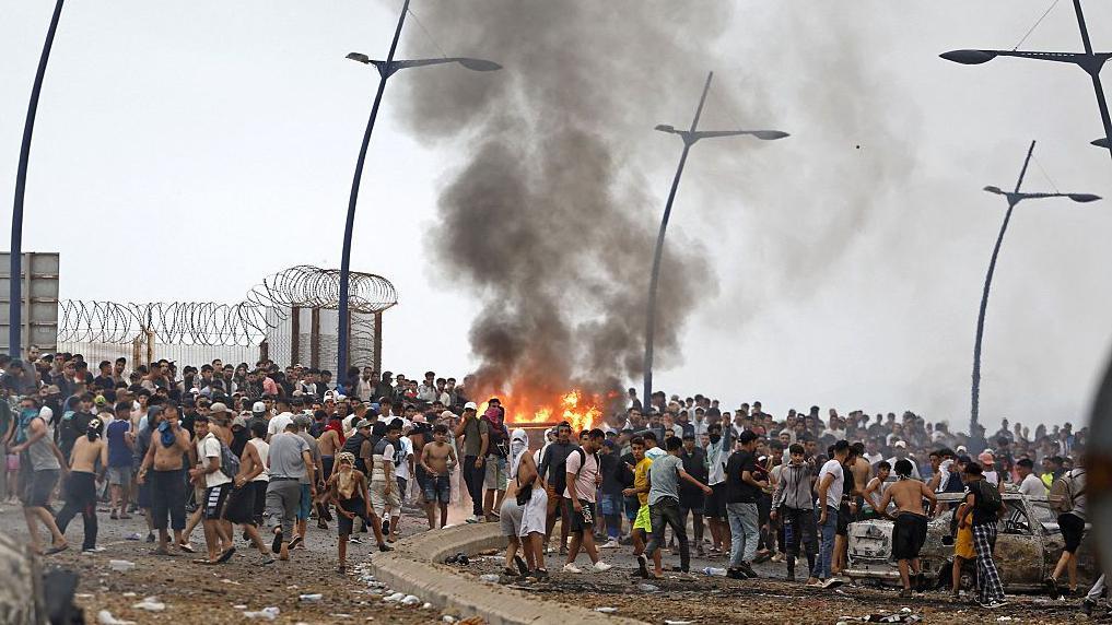 Un fuego y el humo que produce se elevan por el aire en medio de cientos de personas junto a la frontera terrestre en Marruecos y España en Ceuta.