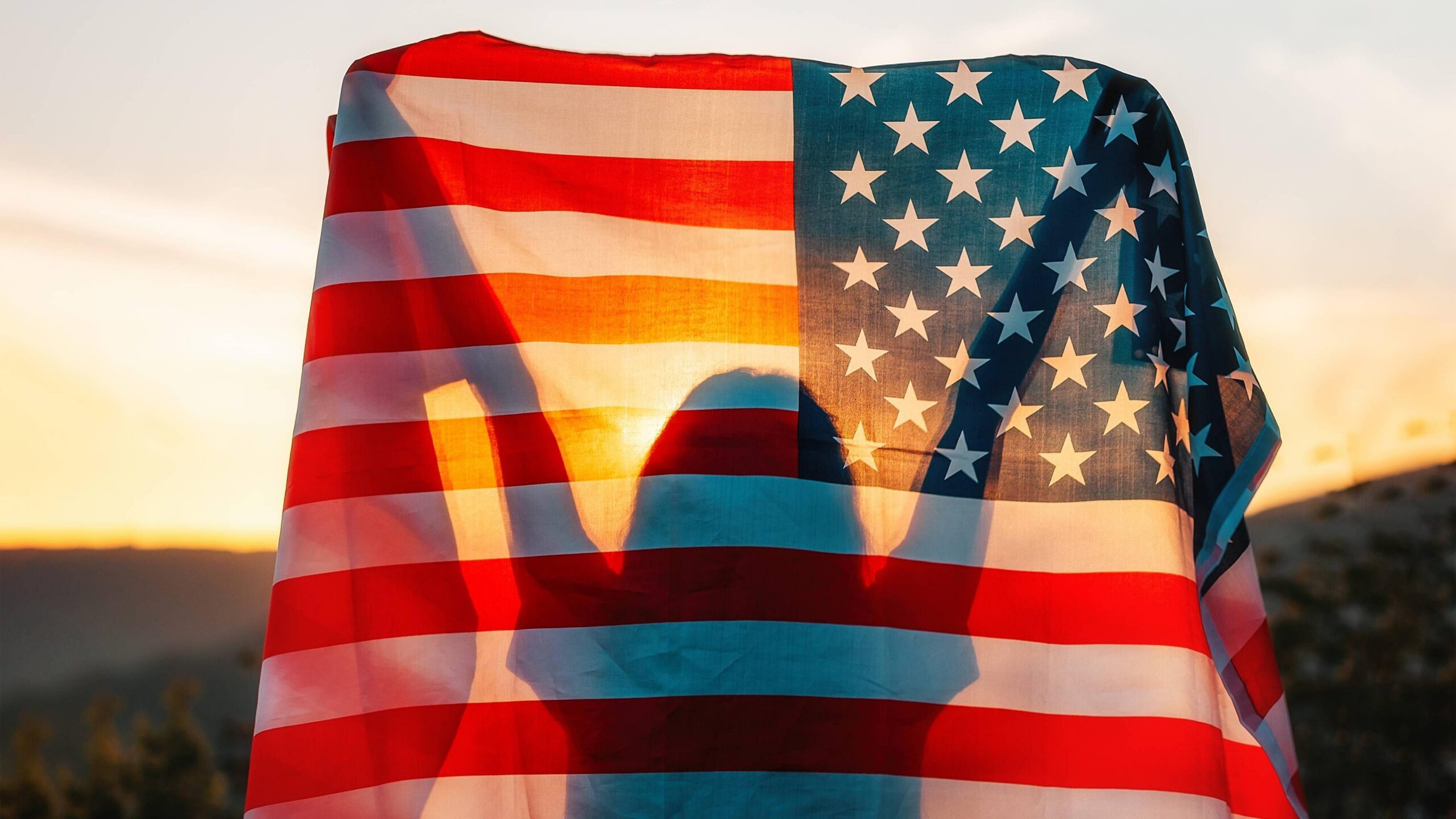 Silueta de una persona con los brazos levantados detr&aacute;s de una bandera de Estados Unidos iluminada por la luz del atardecer.