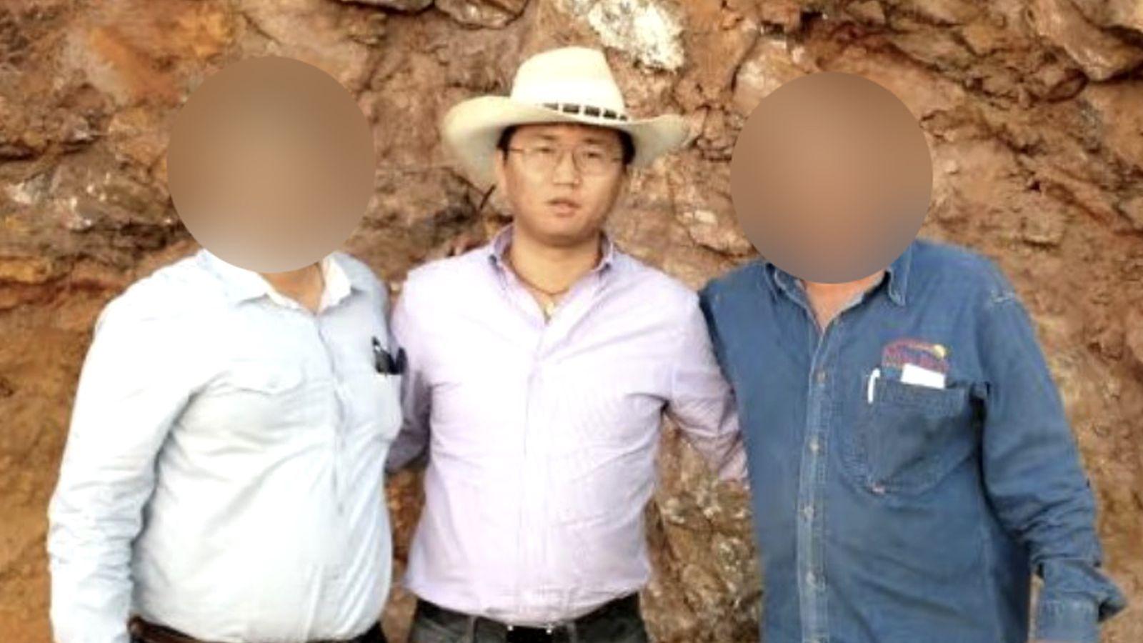 Un hombre lleva un sombrero blanco estilo vaquero y una camisa lila metida por dentro del pantal&oacute;n. Usa gafas y un collar, y mira a la c&aacute;mara. A su izquierda hay un hombre y a su derecha otro; los rostros de ambos aparecen borrosos para ocultar sus identidades. 