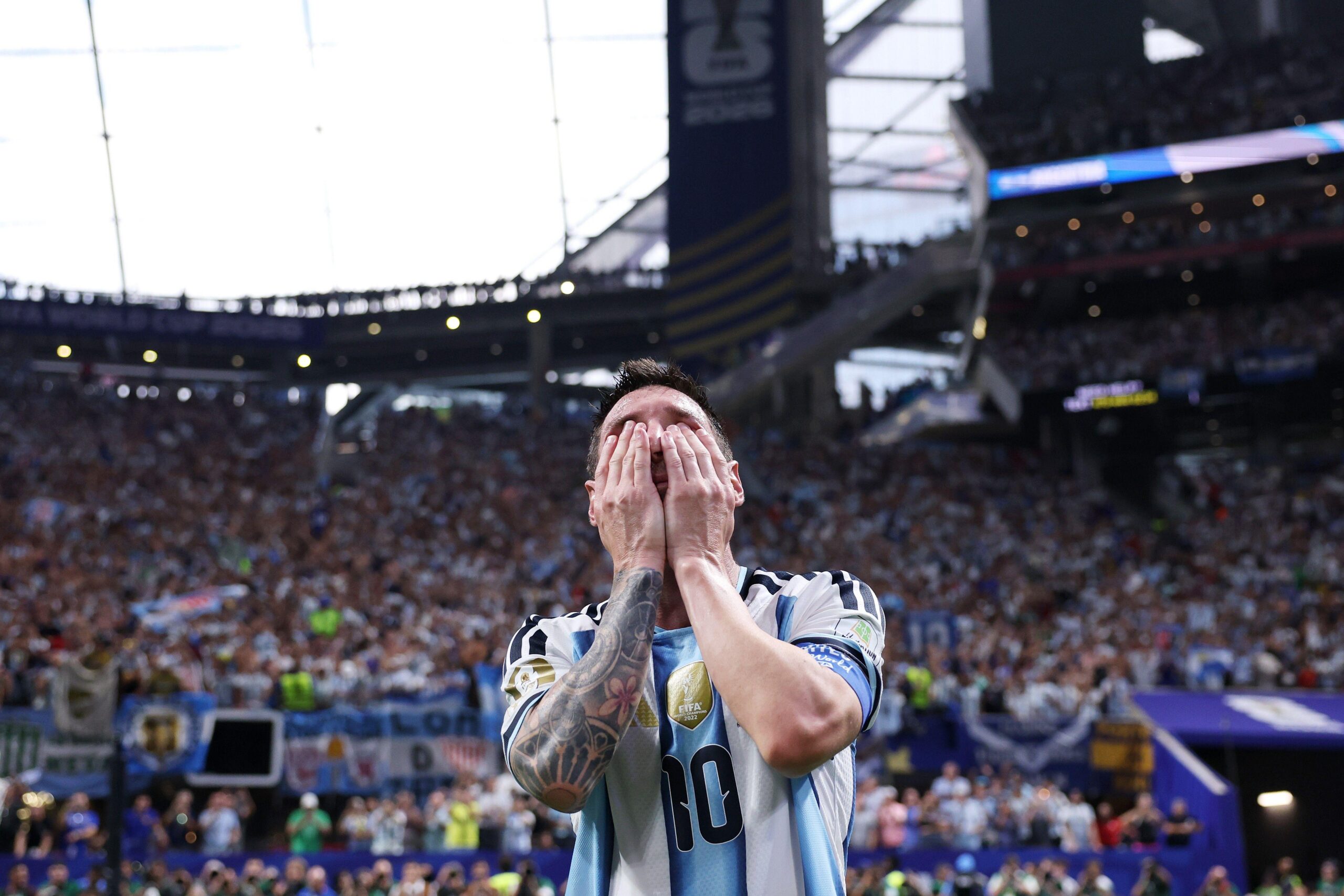 Messi con las manos sobre su rostro, y una tribuna del estadio repleta de aficionados detr&aacute;s.