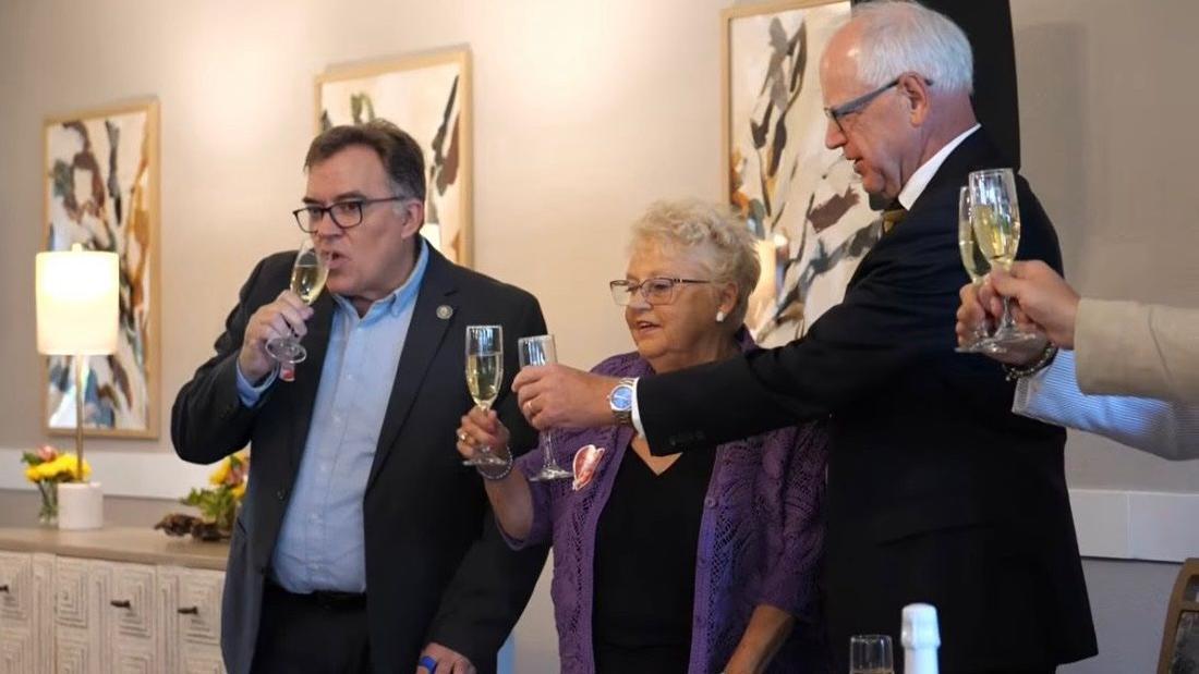 Anita LeBrun, con una chaqueta morada y gafas, alza una copa de champán para brindar con el gobernador de Minnesota, Tim Walz.