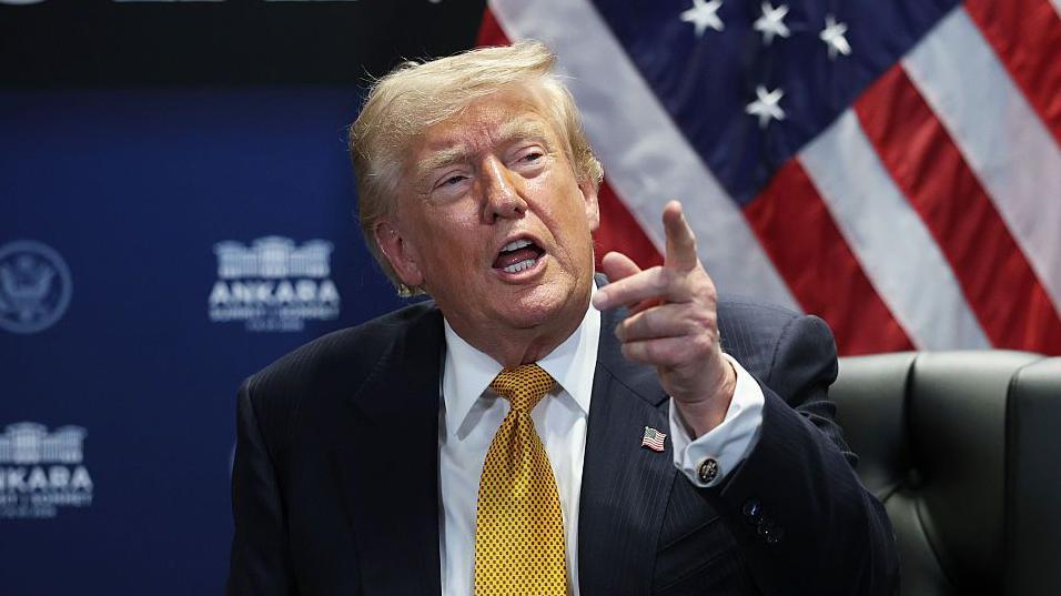 Donald Trump hablando y se&ntilde;alando con el dedo. Viste traje oscuro, corbata en tono amarillo-dorado y camisa blanca.