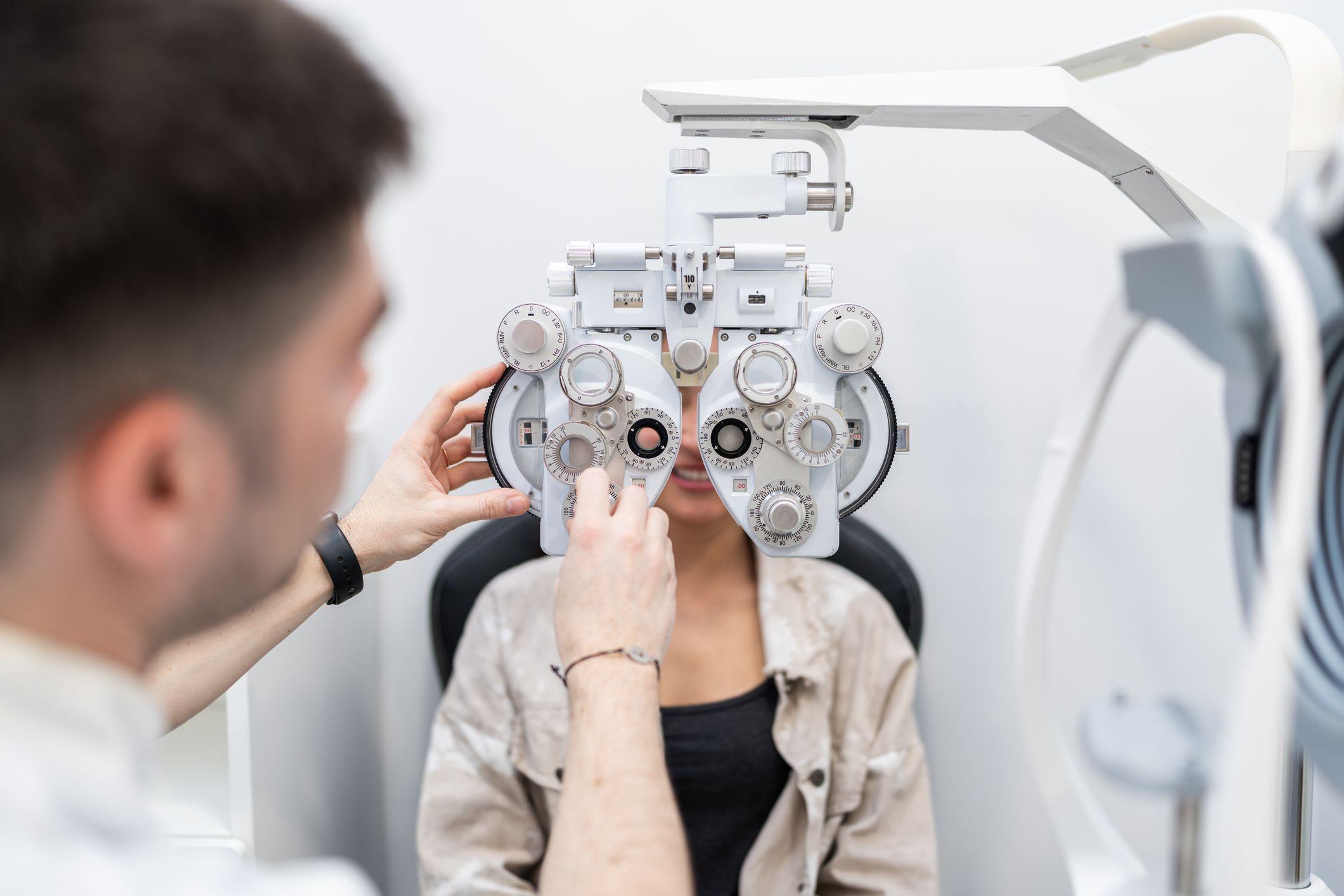 Una mujer est&aacute; sentada en la silla del oftalm&oacute;logo y &eacute;l le tiene el aparato para probar lentes en frente.