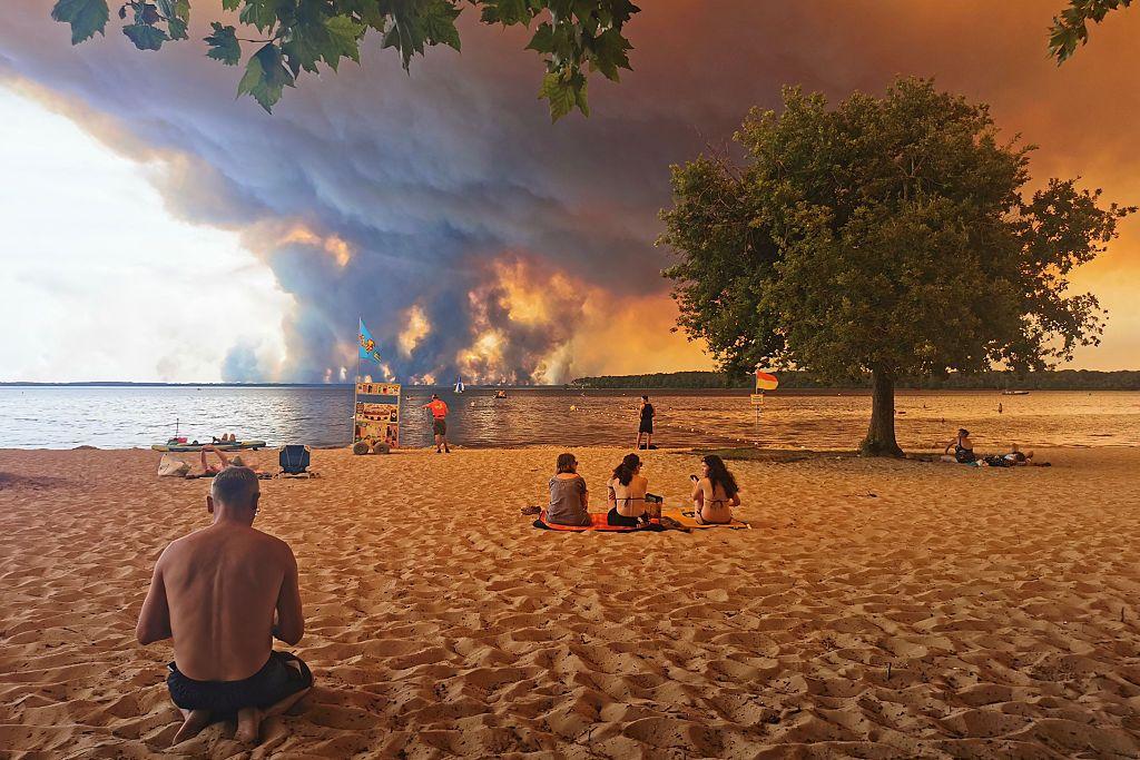 Bañistas descansan en la playa de Moutchic, a orillas del lago Lacanau (Étang de Lacanau), en Lacanau, en el suroeste de Francia, el 24 de julio de 2026, mientras columnas de humo se elevan en el cielo por el incendio forestal de Gironda. 