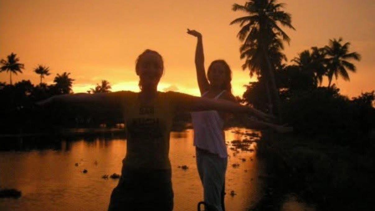 Lowri y una amiga bailando al atardecer en India. Estaban de pie frente a un lago con palmeras en sus orillas.