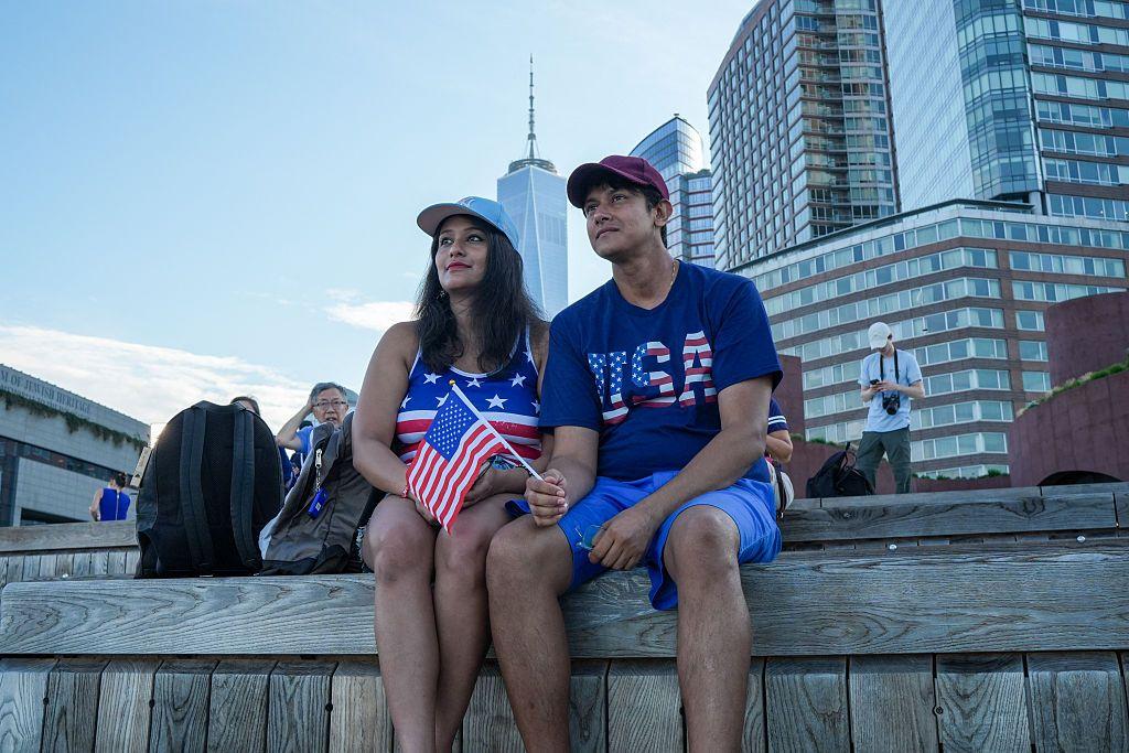 Una pareja observa desde un muelle el desfile de barcos Sail4th 250 en el puerto de Nueva York, con motivo del 250 aniversario de la Independencia de Estados Unidos, el 4 de julio de 2026. Ambos van vestidos con camisetas que llevan la bandera de Estados Unidos, llevan gorras. Uno de ellos lleva una bandera de Estados Unidos en la mano. 
