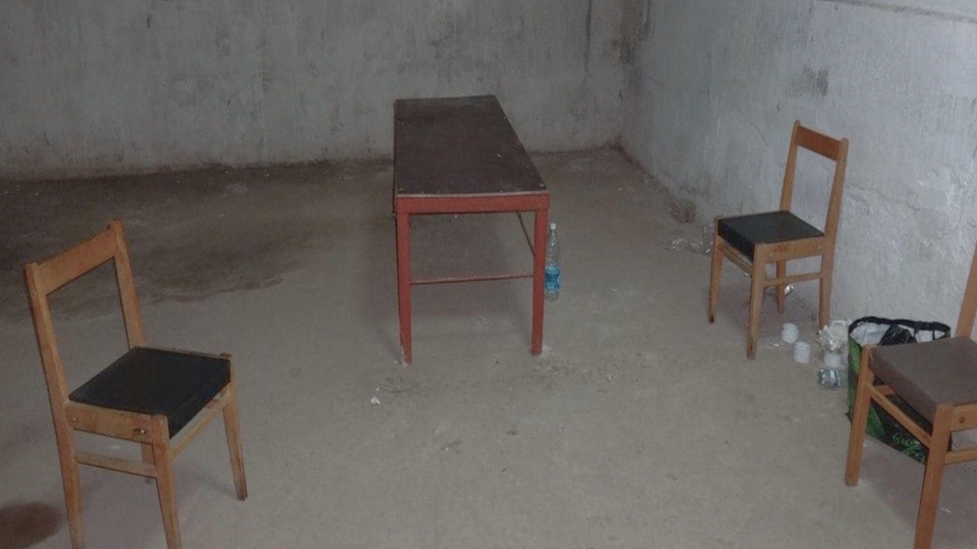 Una habitaci&oacute;n desnuda, con paredes y suelo de hormig&oacute;n. En ella hay tres sillas de madera y una peque&ntilde;a mesa de madera, con unas cuantas botellas de agua de pl&aacute;stico en el suelo.