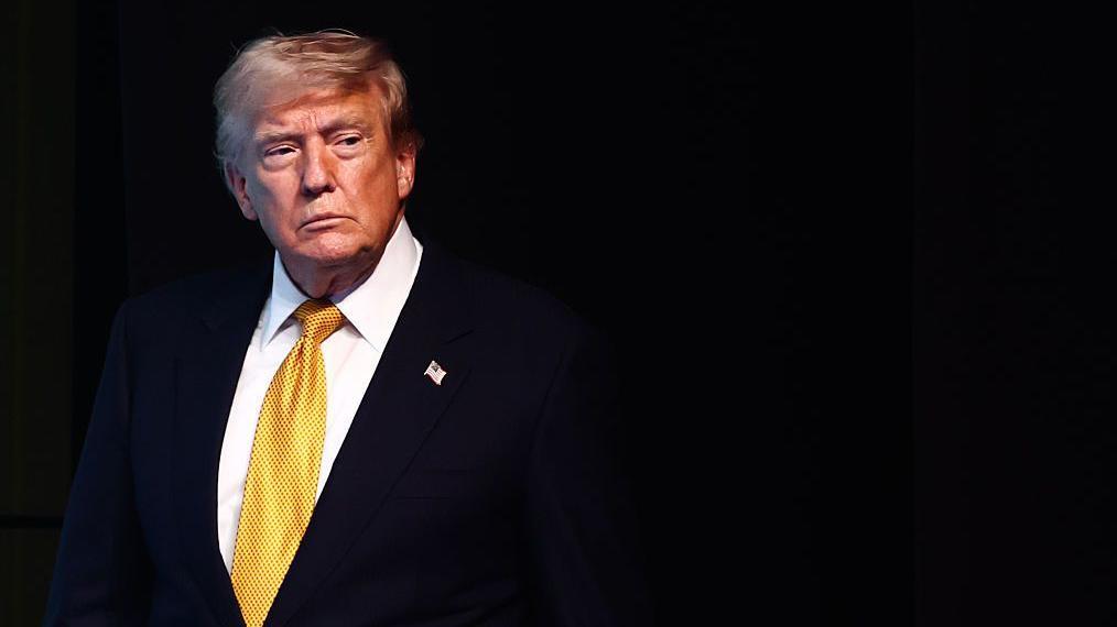 Trump vistiendo un traje con una corbata amarilla. El fondo es negro.