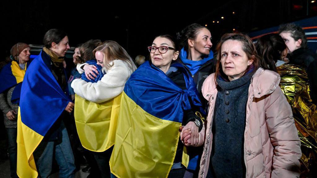 Varias mujeres envueltas en banderas ucranianas azules y amarillas. Dos mujeres se abrazan al fondo y otras sonr&iacute;en. Liudmyla mira hacia su derecha mientras sostiene la mano de una mujer con abrigo rosa situada a su izquierda.