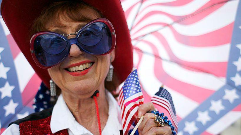 Una mujer con una sombrilla con los colores de la bandera de Estados Unidos, vestida con un sombrero estilo vaquero rojo, chaleco de lentejuelas rojo y azul, camisa blanca y unos lentes azules y rojos. Lleva en su mano dos peque&ntilde;as banderas de Estados Unidos. 