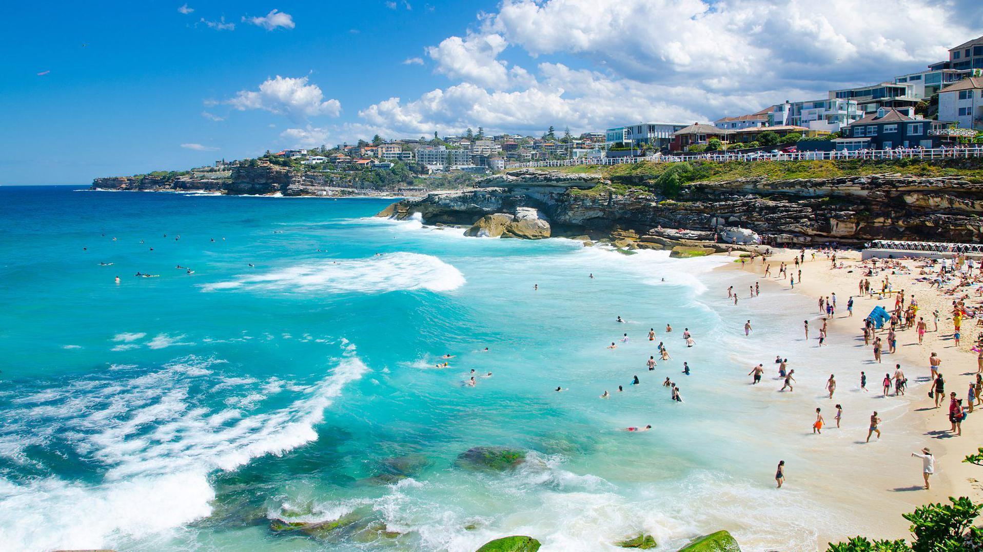 Playa de S&iacute;dney con aguas azules y cristalinas, la gente disfrura adentro y fuera del mar.