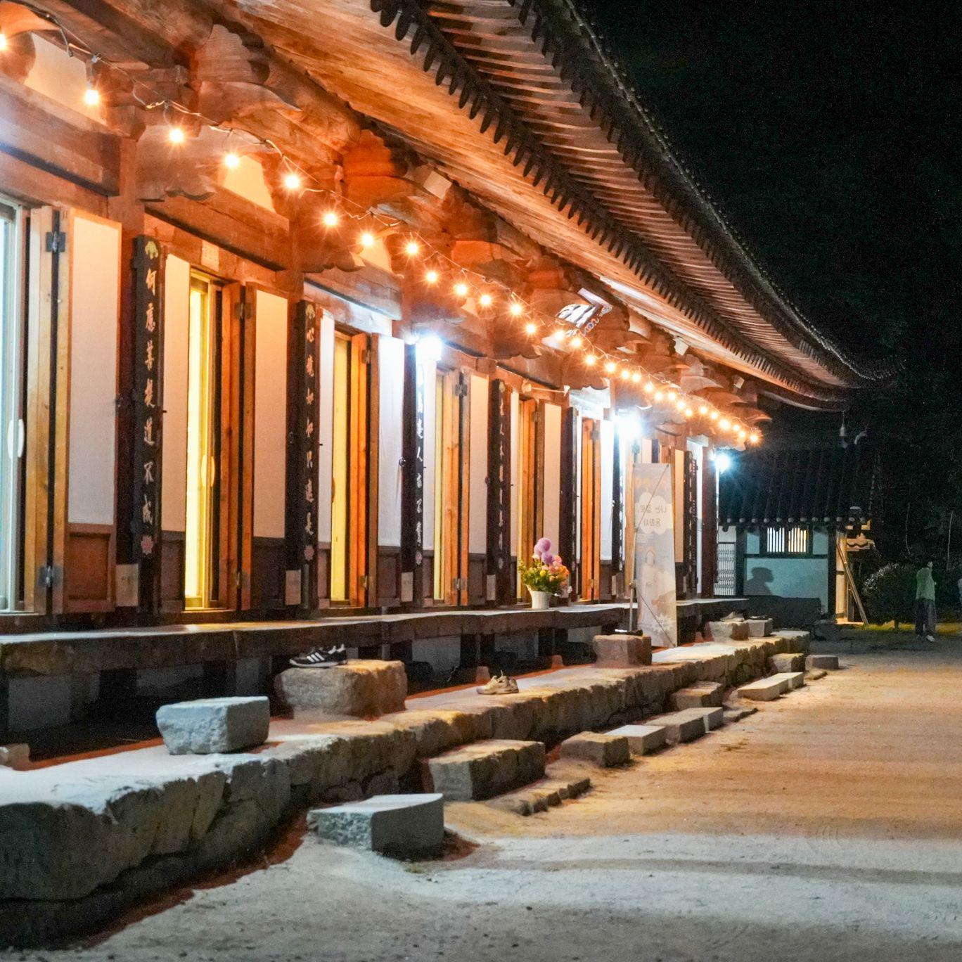 Un templo coreano tradicional iluminado por c&aacute;lidas luces por la noche, con un tejado curvo de tejas, columnas de madera y escalones de piedra que bordean un amplio patio. Varios visitantes caminan por el sendero que discurre junto al templo.