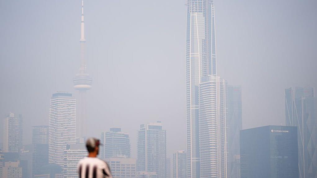 Un hombre con gorra y camisa de rayas verticales blancas y negras contempla el horizonte brumoso de Toronto.