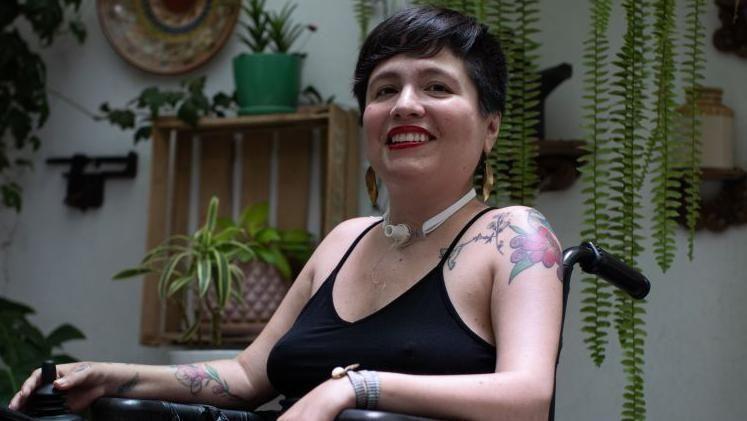 Ana, en primer plano, sentada en una silla de ruedas, con un hueco de traqueotom&iacute;a en la garganta. De fondo hay una pared blanca con muchas plantas. 