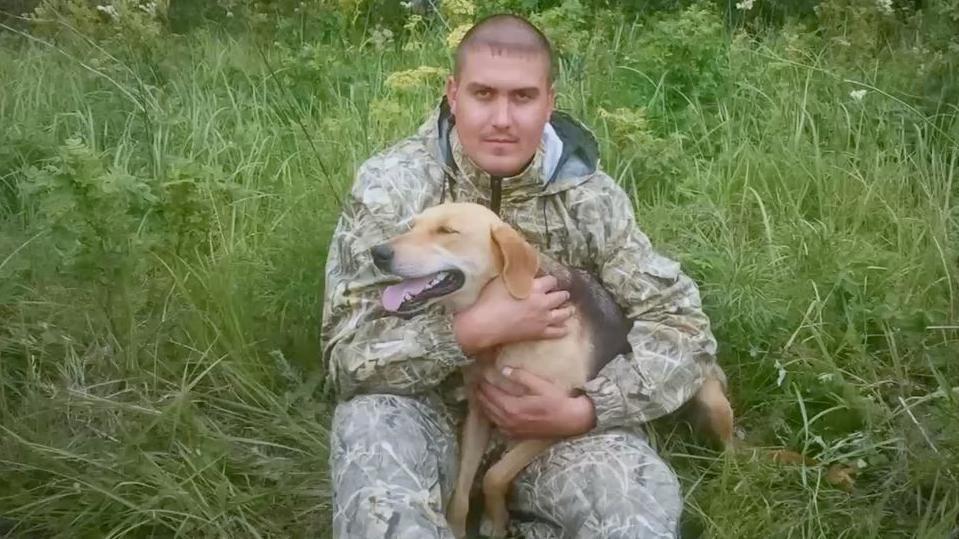 Andrey Spivak, sentado entre hierba alta, sostiene a un perro de color beige y negro. Viste un conjunto de exterior con estampado de camuflaje, lleva el pelo rapado al ras y mira directamente a la c&aacute;mara.